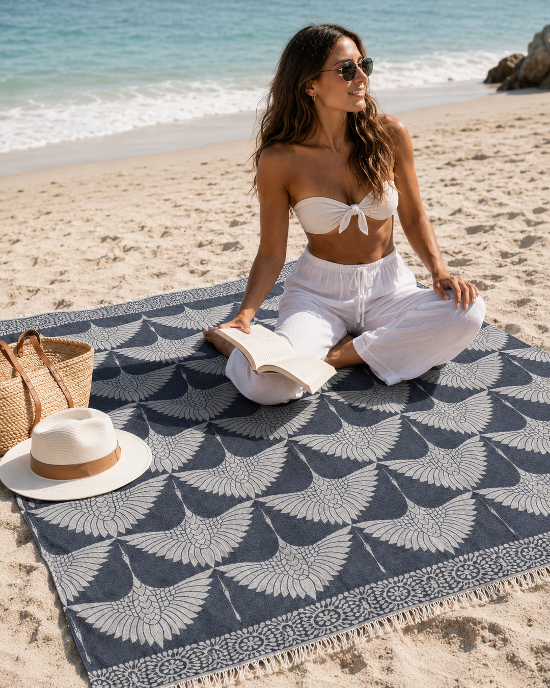 A woman wearing sunglasses, a white bikini top, and white pants sits cross-legged at the beach reading a book on a patterned Turkish muslin cotton blanket. Next to her on the sand near the shoreline are a straw bag and hat, along with the Crane Muslin Throw Blanket Natural Border– Reversible Turkish Cotton by turkanhome.com.