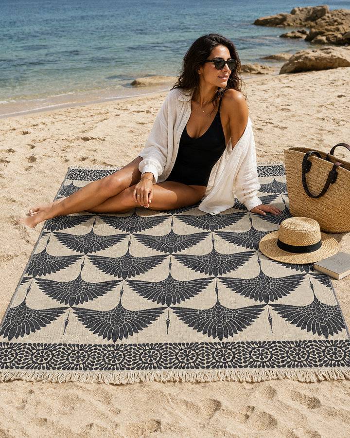 A woman in sunglasses and a black swimsuit sits on a patterned beach blanket by the sea. Beside her on the sand are the Crane Muslin Throw Blanket Natural Border – Reversible Turkish Cotton from turkanhome.com, along with a straw bag, hat, and book. The background features a rocky shoreline and calm blue water.