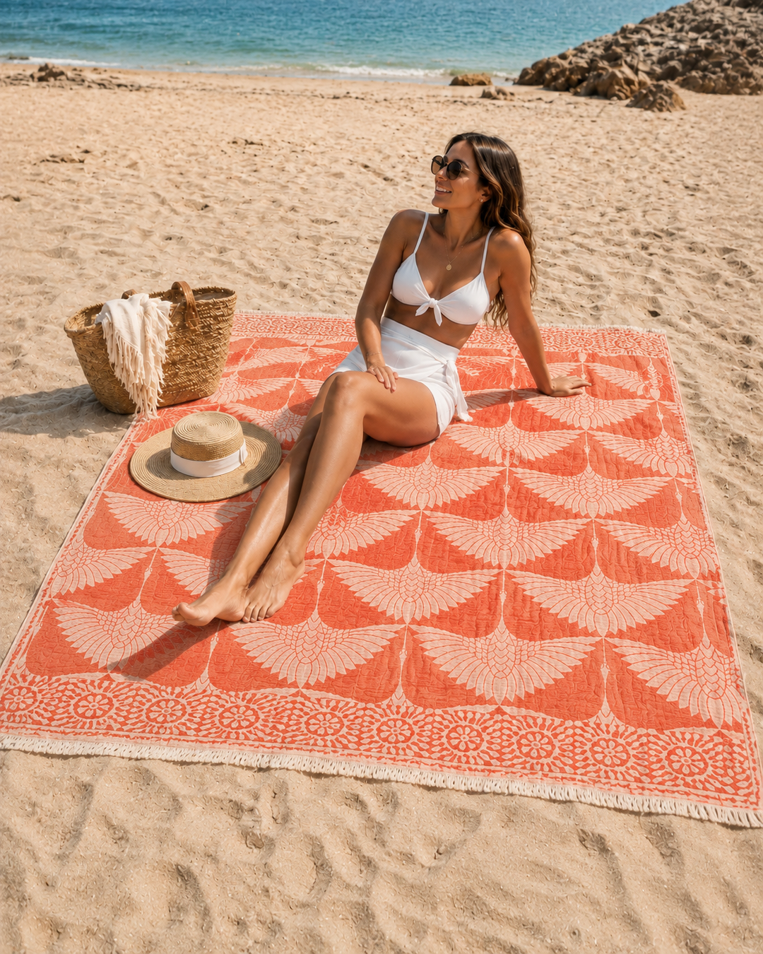 A woman in a white swimsuit relaxes on the sandy shore near the ocean, seated on a Crane Muslin Throw Blanket Natural Border– Reversible Turkish Cotton from turkanhome.com, with a straw hat and woven beach bag placed beside her.