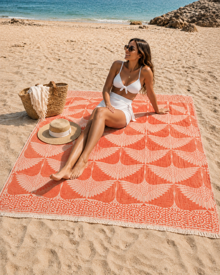 A woman in a white swimsuit relaxes on the sandy shore near the ocean, seated on a Crane Muslin Throw Blanket Natural Border– Reversible Turkish Cotton from turkanhome.com, with a straw hat and woven beach bag placed beside her.