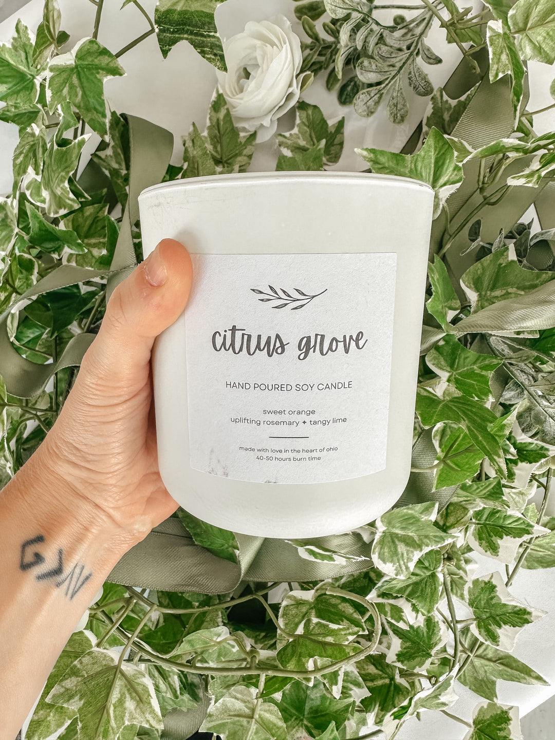 A hand holds the Citrus Grove Beeswax Candle (10oz) by Ampersand Candles + Oils, featuring sweet orange, rosemary, and tangy lime. Surrounded by green plants and a white rose, this all-natural candle brings fresh nature scents to your space.