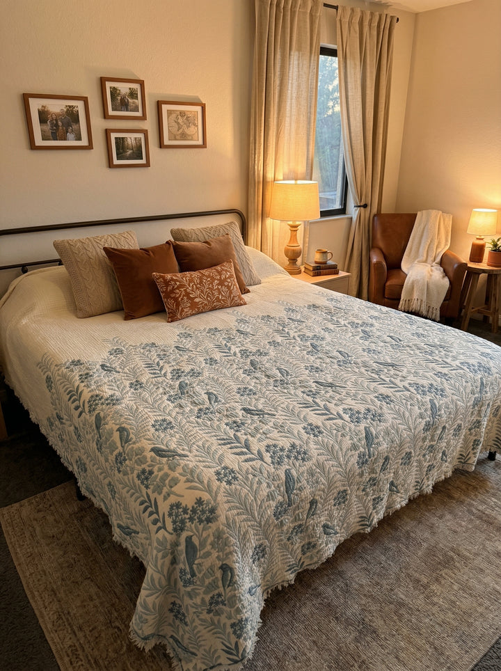 A cozy bedroom features a bed with the Luxury 7-Layer Cotton Muslin Quilted Blanket - Mint Bird by turkanhome.com, decorative pillows, a lamp on a nightstand, framed photos, beige curtains, and a brown armchair with a blanket.