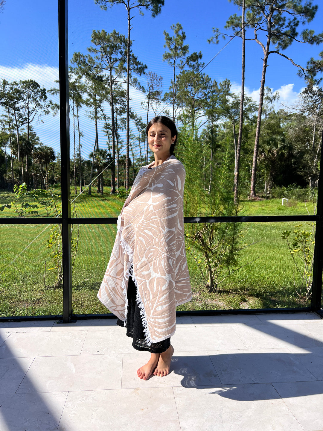 A woman stands on a patio wrapped in a Botanical Turkish Towel from turkanhome.com, with green grass, trees, and a screened enclosure visible beneath a clear blue sky.