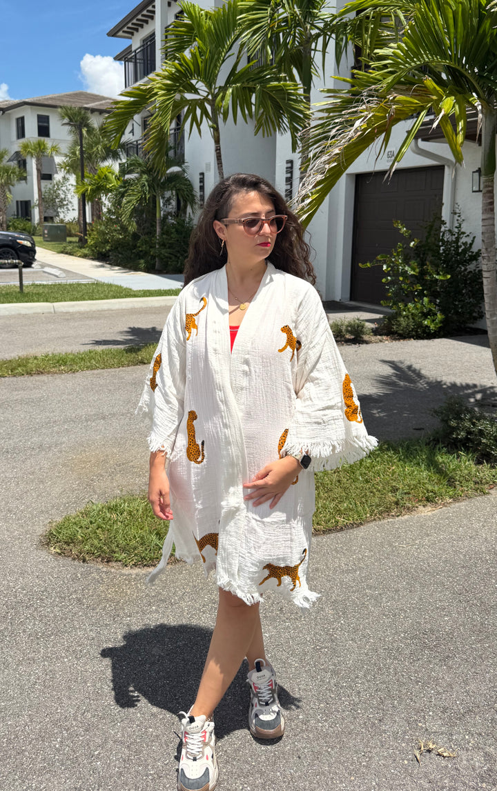 A woman in sunglasses and sneakers wears the Leopard Cotton Kimono Robe-Short from turkanhome.com, featuring a white boho-chic design with leopard prints, over a red top. She stands on a sunny suburban street lined with palm trees and houses.