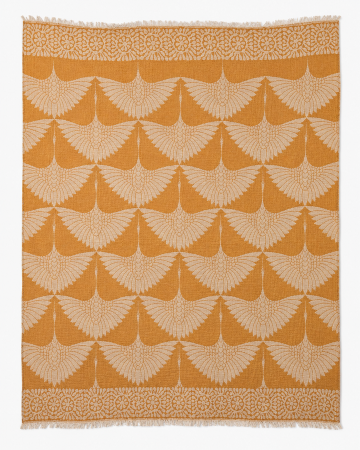 Introducing the Crane Muslin Throw Blanket Natural Border–Reversible Turkish Cotton by turkanhome.com: a square throw blanket crafted from premium Turkish cotton, featuring a repeating pattern of cream-colored, stylized cranes with outstretched wings set against a rich mustard yellow background. Accented with an elegant natural-hued geometric border, this reversible blanket effortlessly adds both sophistication and comfort to your living space.