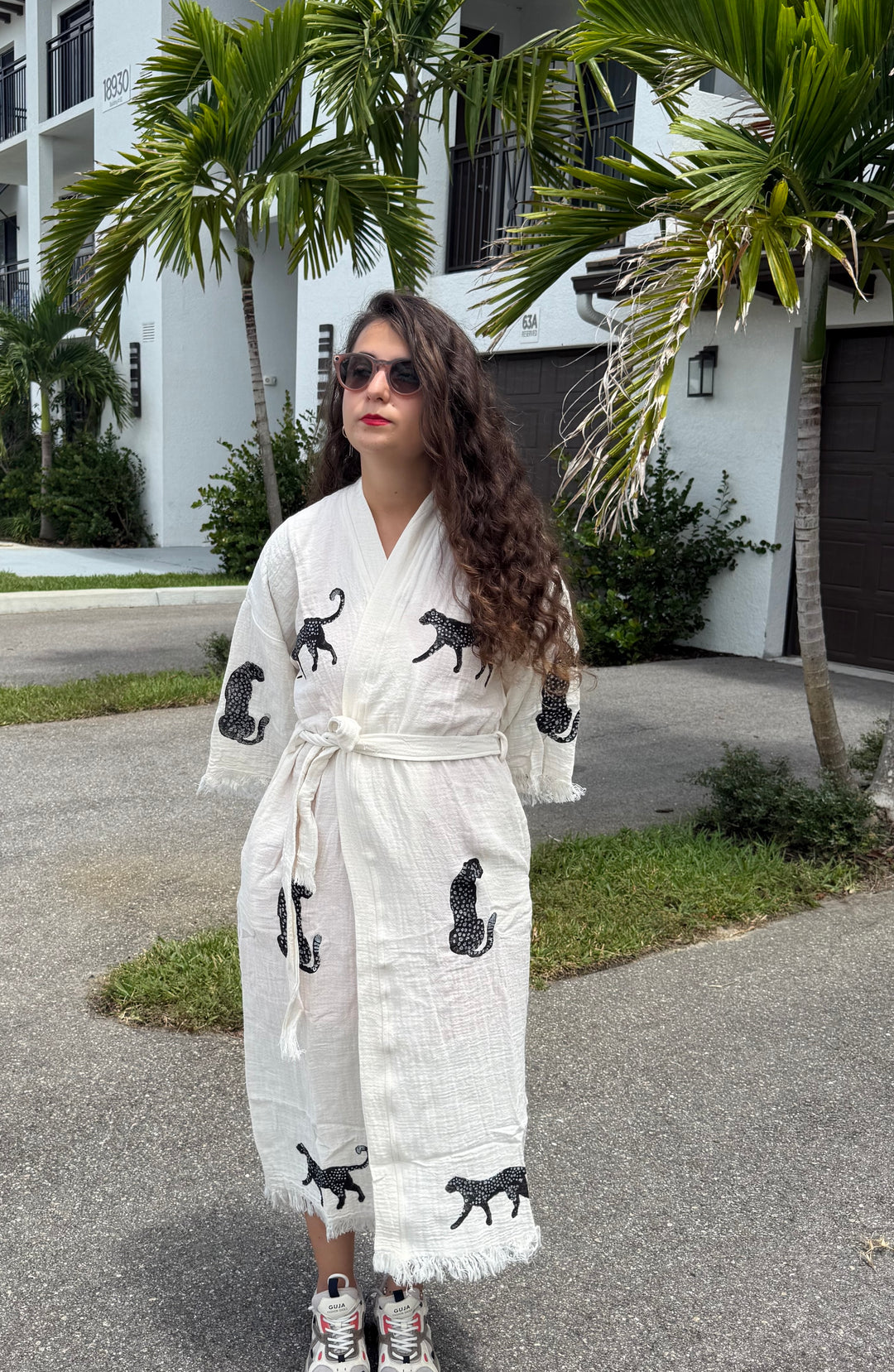A woman with long curly hair, wearing sunglasses and a stylish Leopard Cotton Kimono Robe – Lightweight Boho Beach Cover Up by turkanhome.com featuring black leopard prints, stands on a driveway near palm trees and a white building.