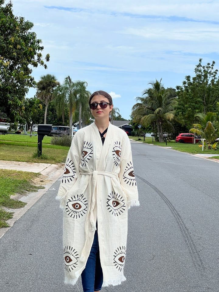 A woman in a Brown Eye Kimono Robe by turkanhome.com and sunglasses strolls down a sunny, palm-lined suburban street with parked cars.
