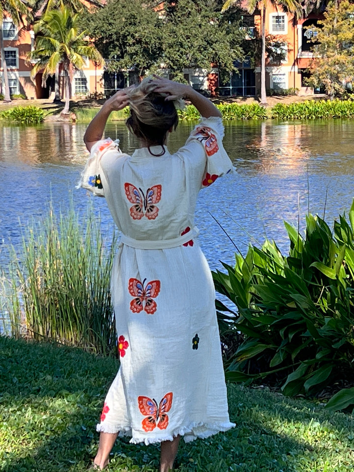 A woman wearing the Butterfly Kimono Robe-Natural Color by turkanhome.com, featuring colorful butterfly and flower prints, stands by a pond with her back turned and hands on her head. Lush plants, trees, and buildings are in the background.