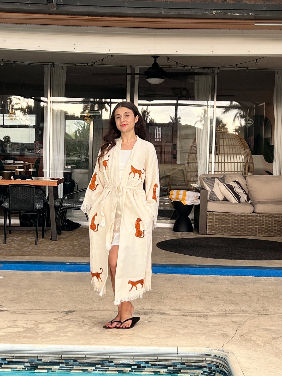 A woman in an Orange Leopard Cotton Kimono Robe by turkanhome.com stands poolside near modern patio furniture and glass doors reflecting palm trees.