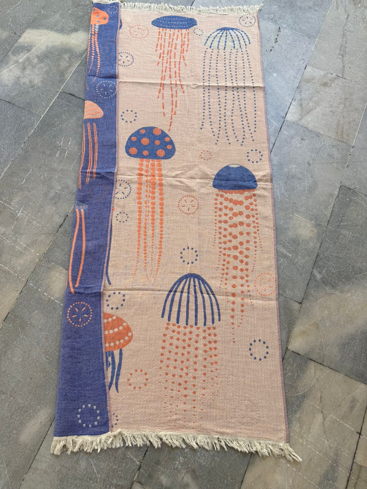 The Jellyfish Reversible Turkish Beach Towel by turkanhome.com is beige with orange and blue jellyfish and bubbles, bordered by a blue strip. Quick drying, sand repellant, and finished with short white fringe at both ends.
