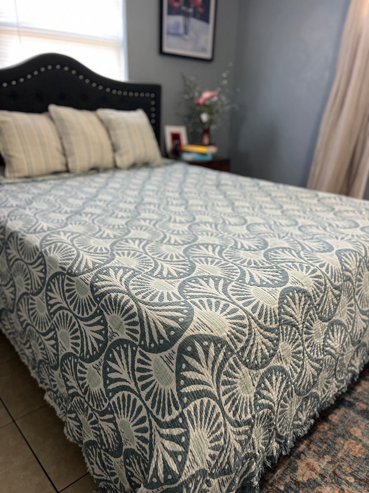 A neatly made bed features the Gingko Muslin Bed Blanket in Teal (King-Oversized Queen) from turkanhome.com, a patterned blue and white bedspread, three striped pillows, and a dark headboard. A window, picture, and nightstand with flowers are in the background.