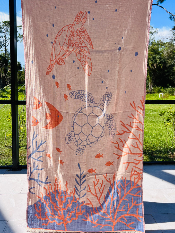The Reef and Turtle Towel – Quick Drying Towel by turkanhome.com displays sea turtles, orange coral, seaweed, and small fish on a light background, hanging vertically with a sunny garden in the background.
