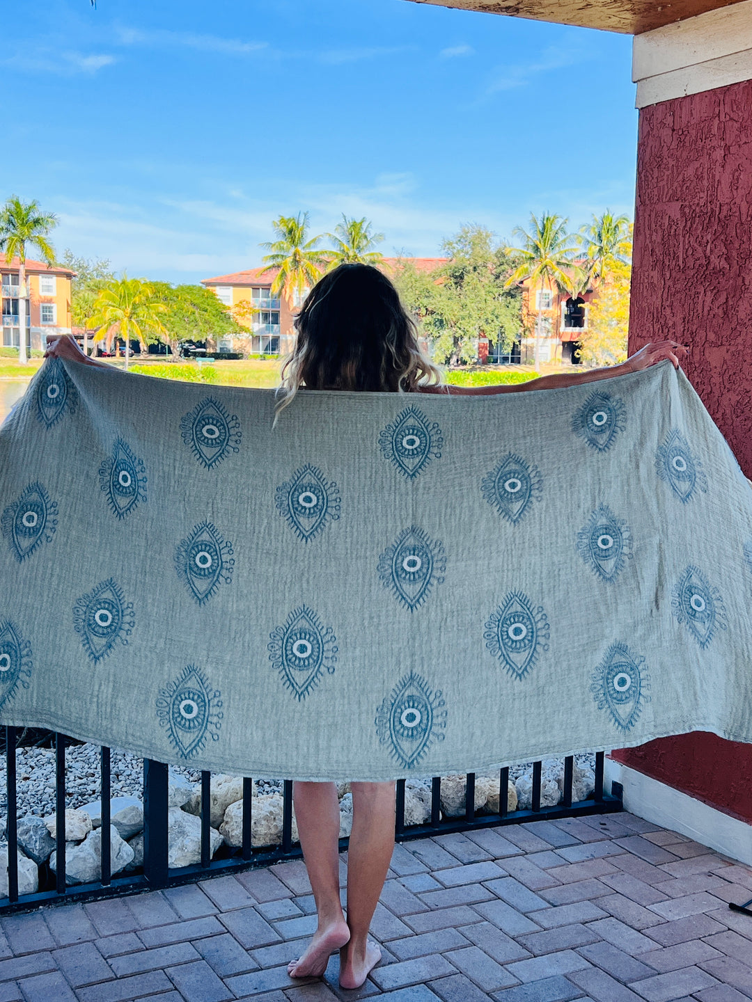 A person with wavy hair stands on a patio holding out the Eye Sage Sand Resistant Turkish Beach Towel by turkanhome.com, featuring blue geometric patterns. Palm trees and buildings appear in the background under a blue sky.