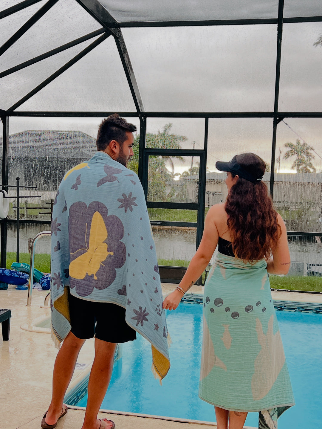 Two people with towels stand by an indoor pool. The person on the left has a turkanhome.com Jacquard Woven towel with a large yellow butterfly, while the person on the right wears a visor and a Dolphin and Whale Turkish Towel.
