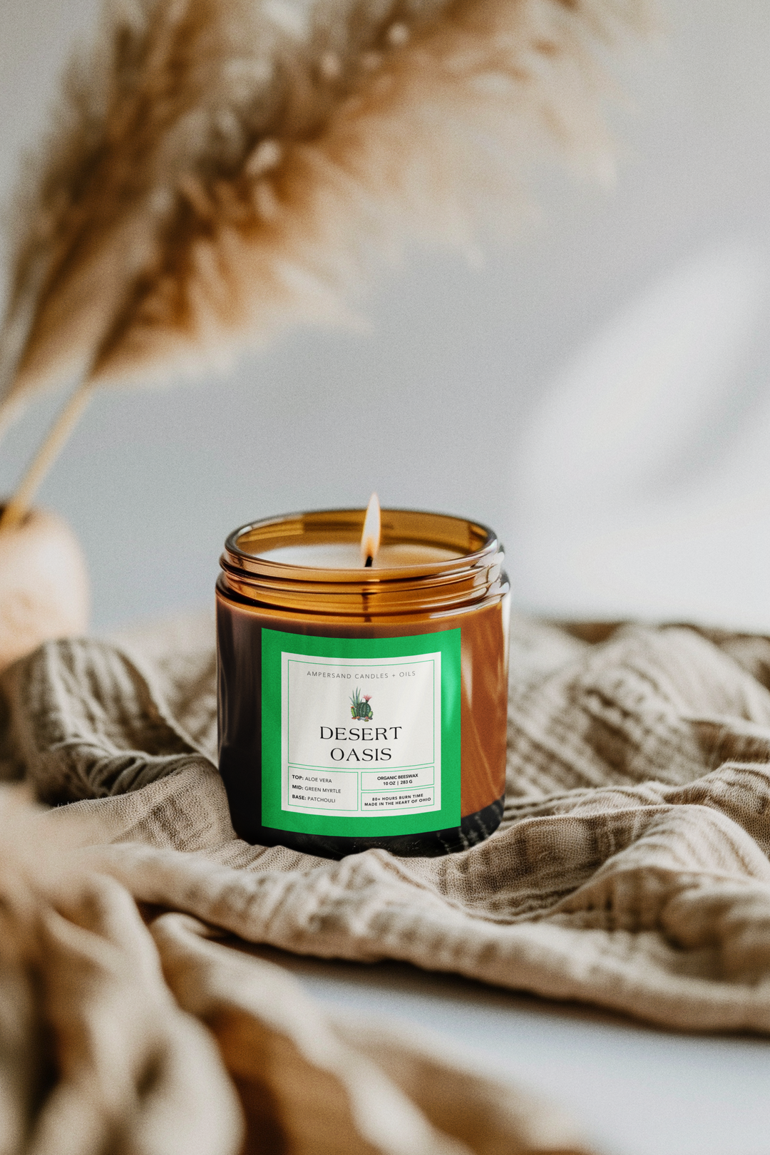 The 10oz Desert Oasis Beeswax Candle by Ampersand Candles + Oils sits on textured beige fabric, with soft, blurred pampas grass behind it, filling the room with a natural aroma.