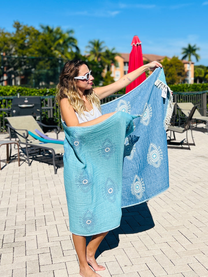 A woman stands barefoot by a pool holding two Eye Blue Sand Resistant Turkish Beach Towels from turkanhome.com—one blue and one turquoise—wearing sunglasses and wrapped in a jacquard woven towel, with lounge chairs and palm trees behind her.