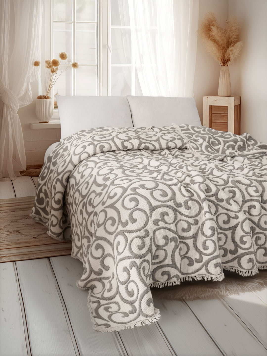 A bright room with a wooden floor features the Serenity Swirl Muslin Quilt— a gray-and-white 5-layer comforter by turkanhome.com—on the bed with white pillows, white curtains, and vases of dried flowers by the window and bedside table.