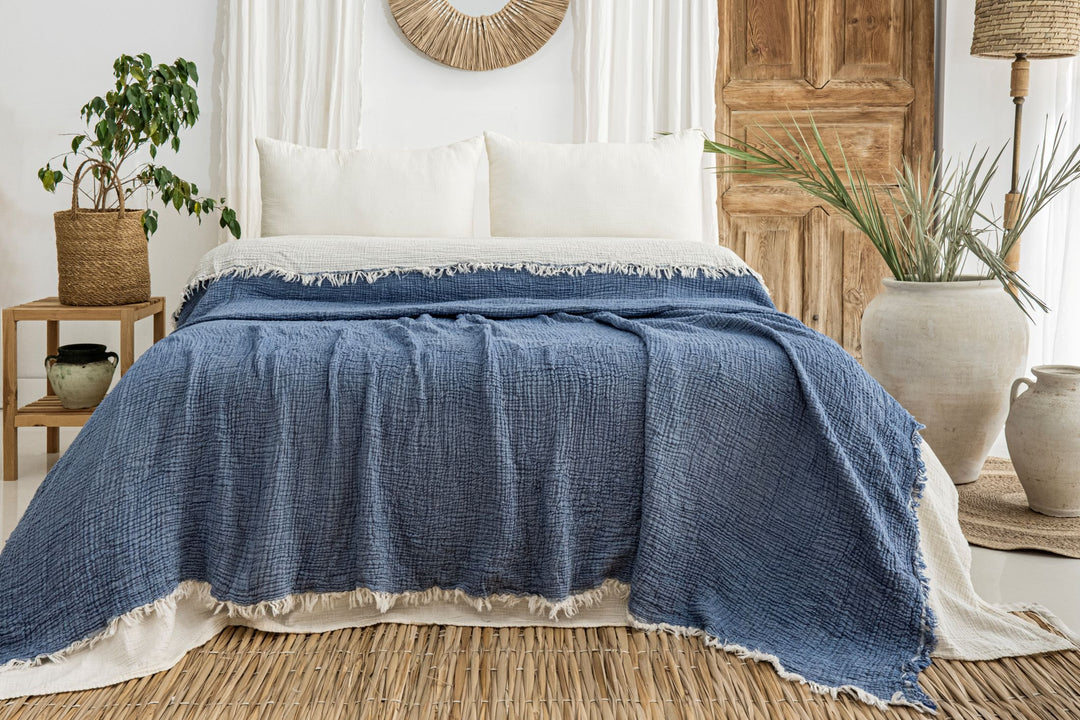 A cozy bed with white pillows and the Indigo Crinkled Muslin Bed Blanket from turkanhome.com sits in a boho-style room with natural wood accents, woven decor, potted plants, and neutral-toned pottery.