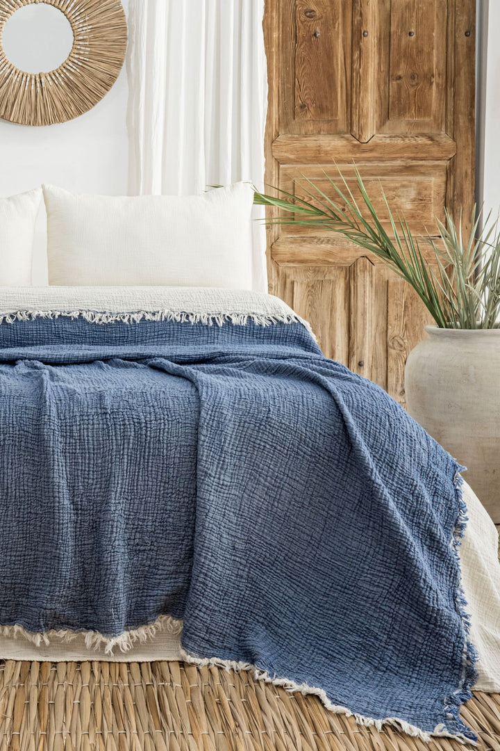 A cozy bed with white pillows and the Indigo Crinkled Muslin Bed Blanket from turkanhome.com sits before a rustic wooden door. In the background are a large planter with green leaves and a round woven-framed mirror.