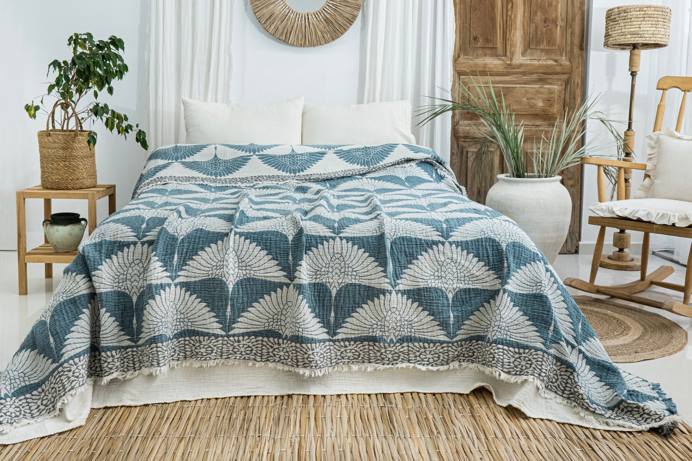 A cozy, light-filled bedroom features the Crane Muslin Bed Blanket Teal from turkanhome.com on a neatly made bed, complemented by white pillows, neutral decor, plants, a rocking chair, wooden door, and a woven rug.
