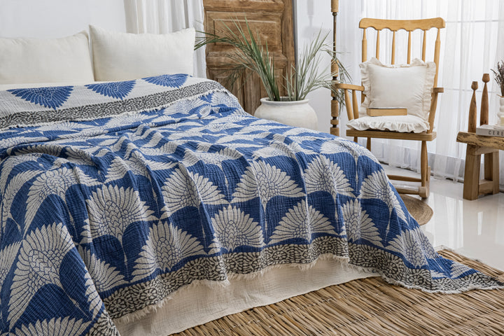 A cozy, bright room features a bed dressed with a Crane Muslin Bed Blanket Blue from turkanhome.com, alongside a wooden rocking chair with cushion, a large white vase of green plants, and airy white curtains.