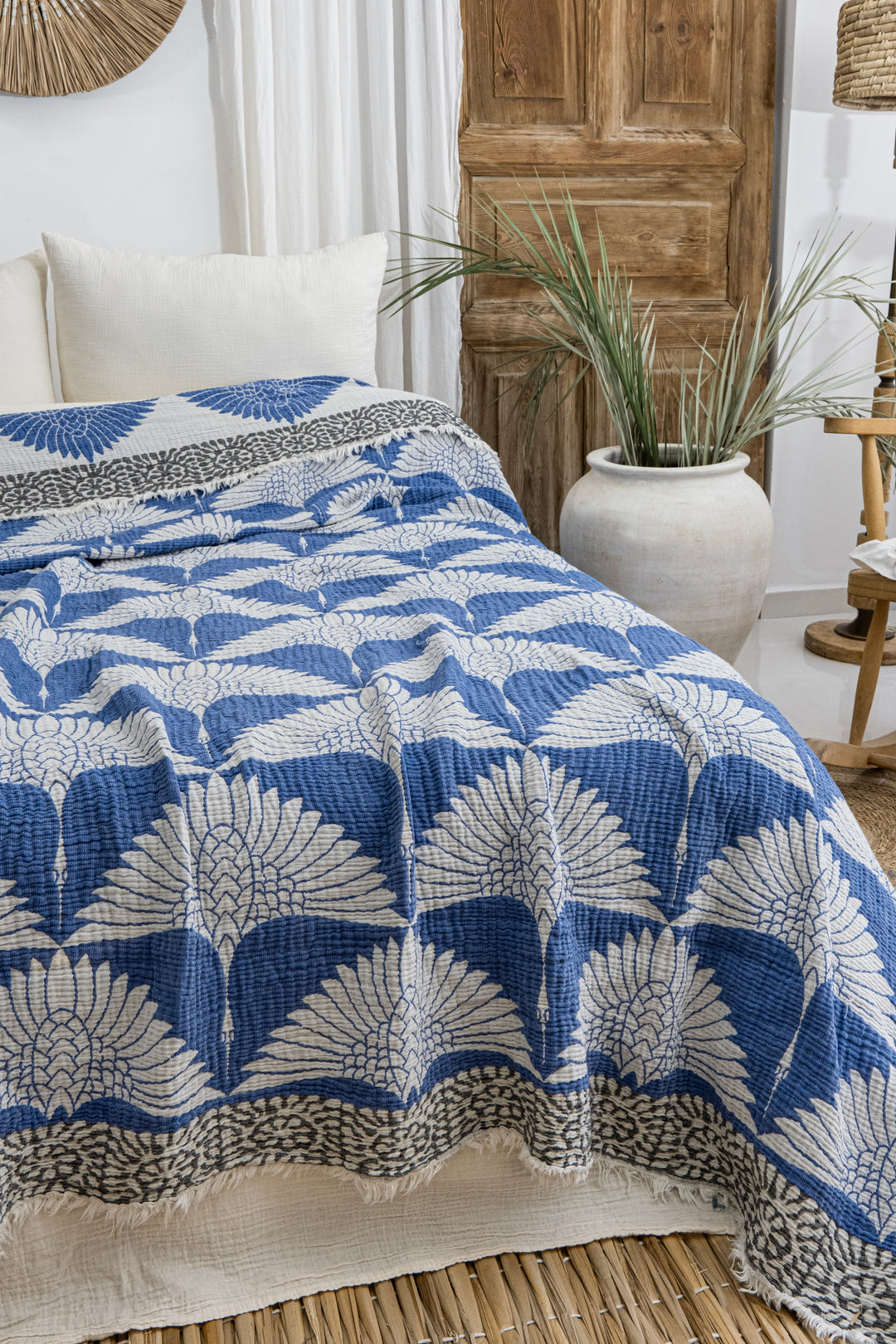 A bed topped with the Crane Muslin Bed Blanket Blue from turkanhome.com and a blue and white leaf-patterned quilt sits in a cozy room with white pillows, a wooden door, greenery in a ceramic vase, and woven accents.