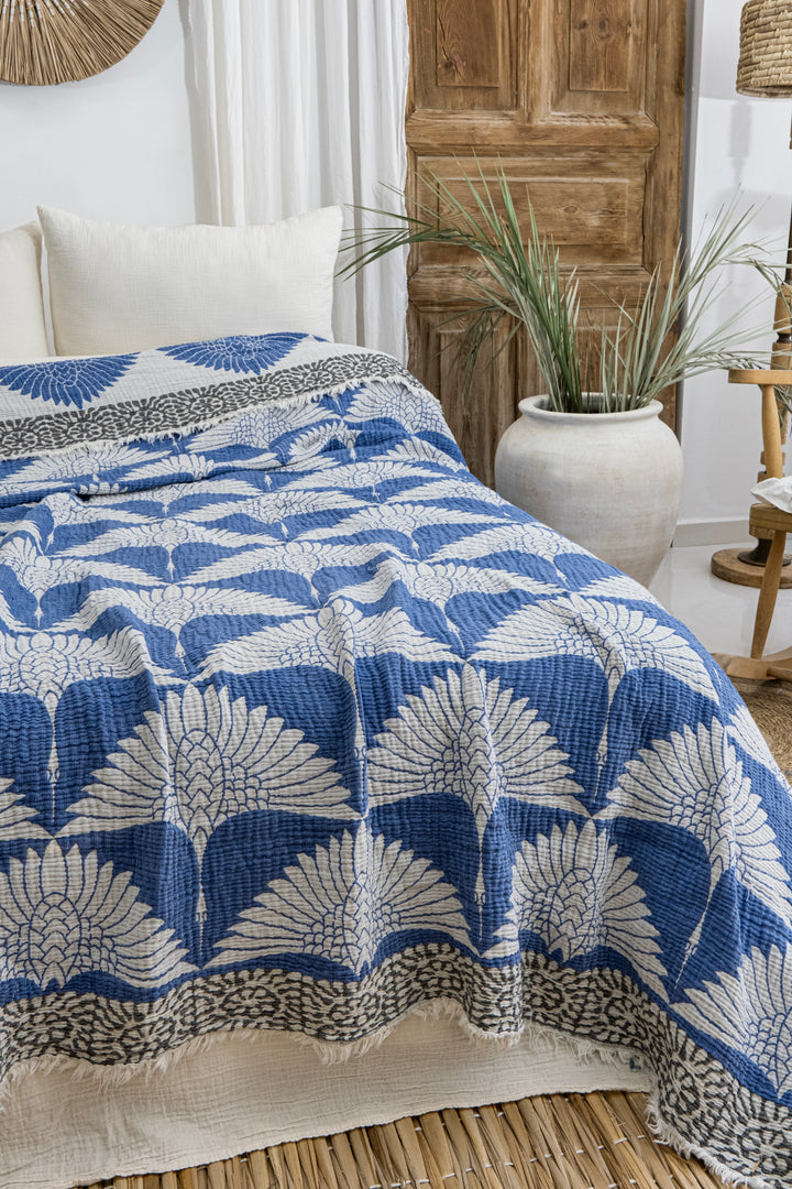 A bed topped with the Crane Muslin Bed Blanket Blue from turkanhome.com and a blue and white leaf-patterned quilt sits in a cozy room with white pillows, a wooden door, greenery in a ceramic vase, and woven accents.