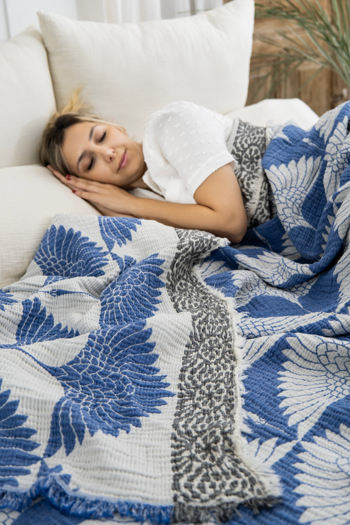 A blonde woman sleeps peacefully on a bed, covered with the Crane Muslin Bed Blanket Blue by turkanhome.com, featuring blue and white patterns, her head resting on a white pillow.