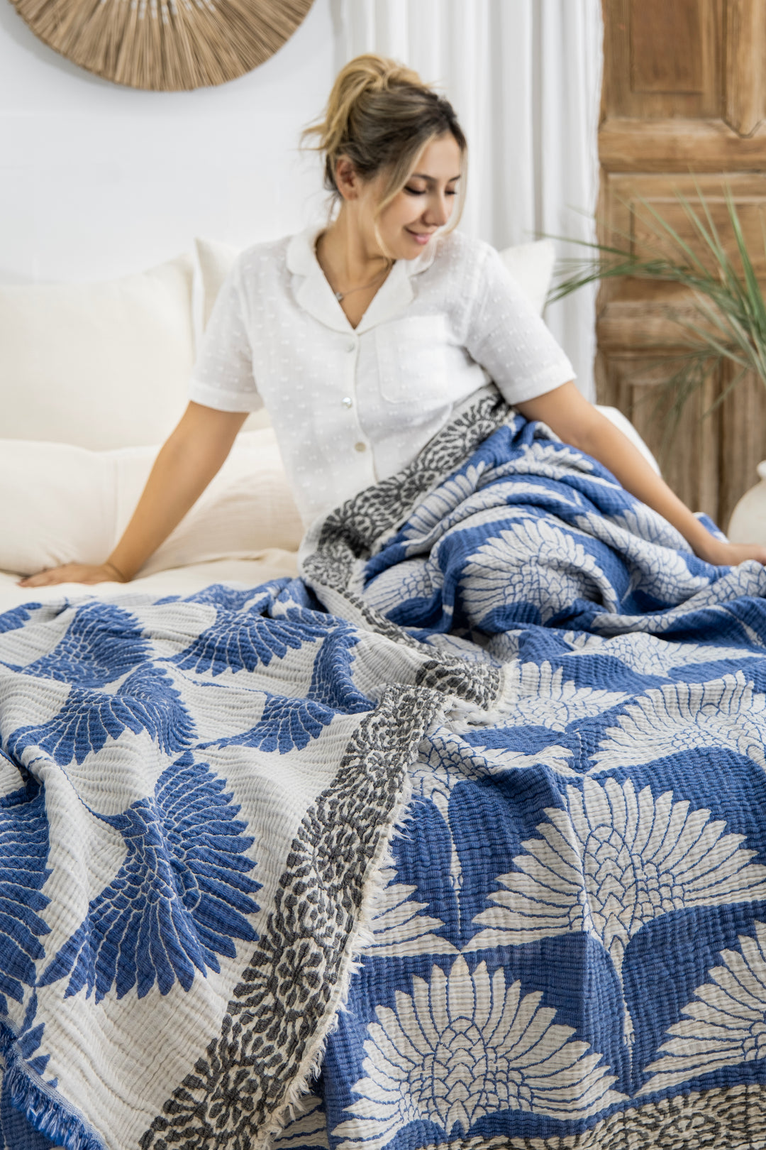 A woman in white pajamas smiles while sitting on a bed with the Crane Muslin Bed Blanket Blue from turkanhome.com, featuring blue and white bird and leaf designs draped over her legs in a cozy, neutral room.