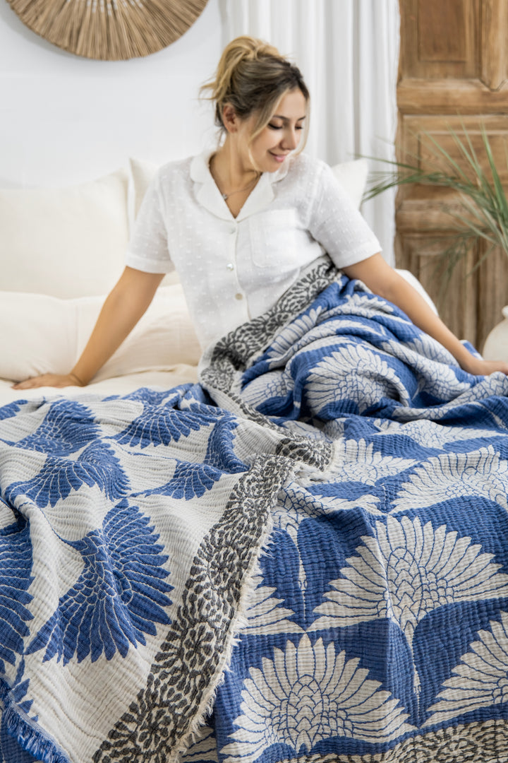 A woman in white pajamas smiles while sitting on a bed with the Crane Muslin Bed Blanket Blue from turkanhome.com, featuring blue and white bird and leaf designs draped over her legs in a cozy, neutral room.