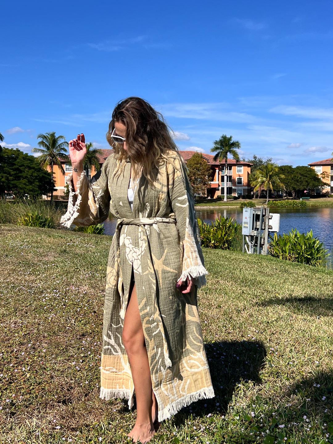A woman in the Green Turtle Robe kimono from turkanhome.com and sunglasses stands barefoot on grass by a pond, with palm trees and colorful buildings in the sunny background.