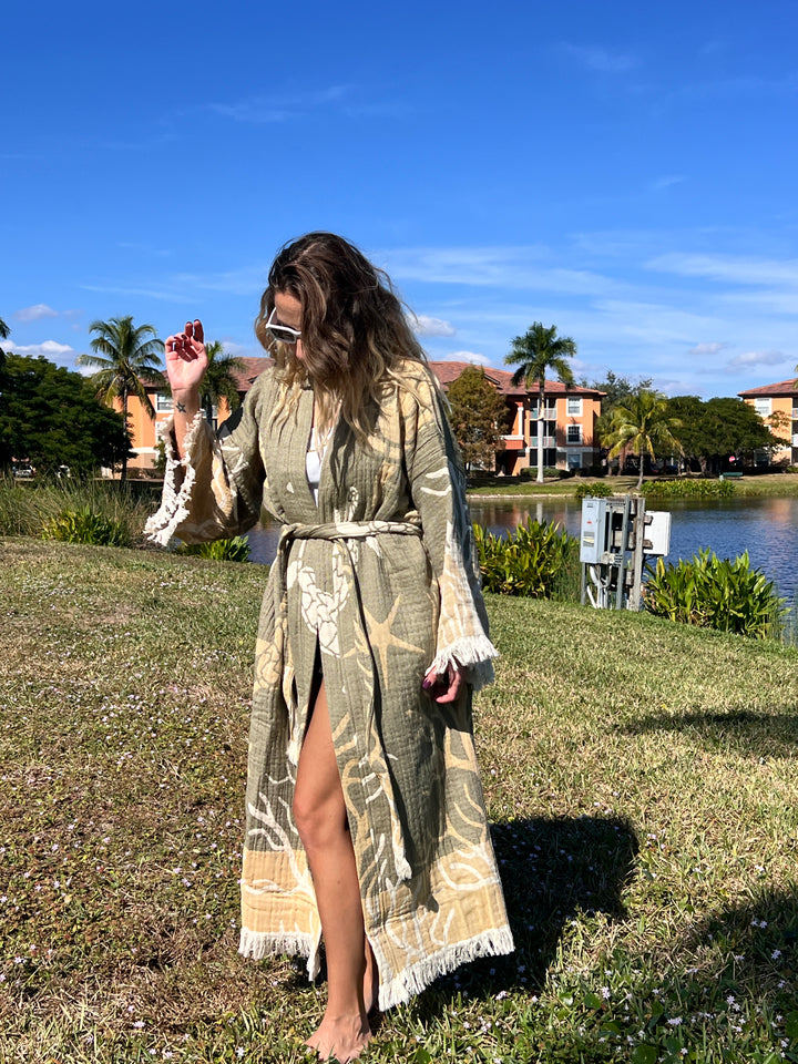 A woman in the Green Turtle Robe kimono from turkanhome.com and sunglasses stands barefoot on grass by a pond, with palm trees and colorful buildings in the sunny background.