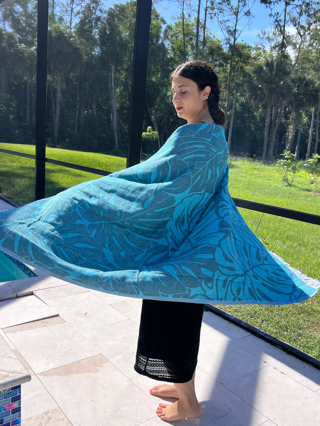 A woman stands barefoot by a pool, wrapped in a blue-leaf-patterned Botanical Turkish Towel from turkanhome.com. She is outdoors on a sunny day, with green grass and tall trees in the background.