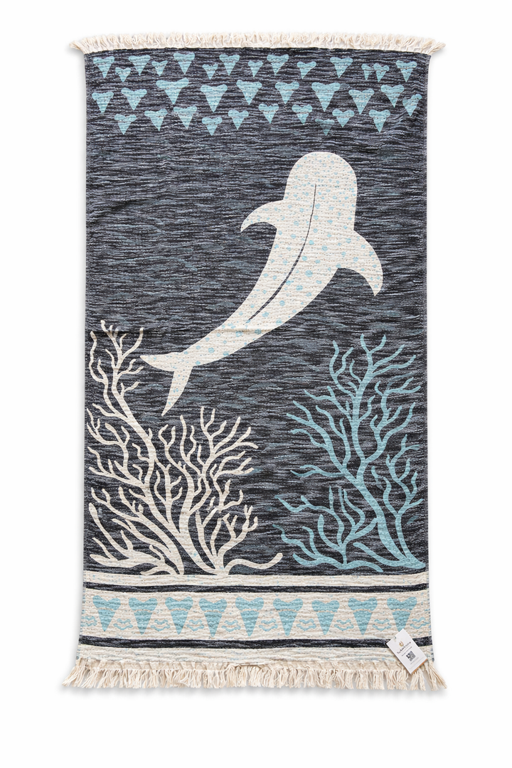 This dark gray woven rug showcases a central white dolphin, accented by light blue and white coral motifs below and geometric blue and white patterns along the top and bottom. Taking inspiration from the intricate designs of turkanhome.com’s Whale Shark Towel – Reversible Turkish Beach Towel, the rug is finished with fringes on its shorter edges.