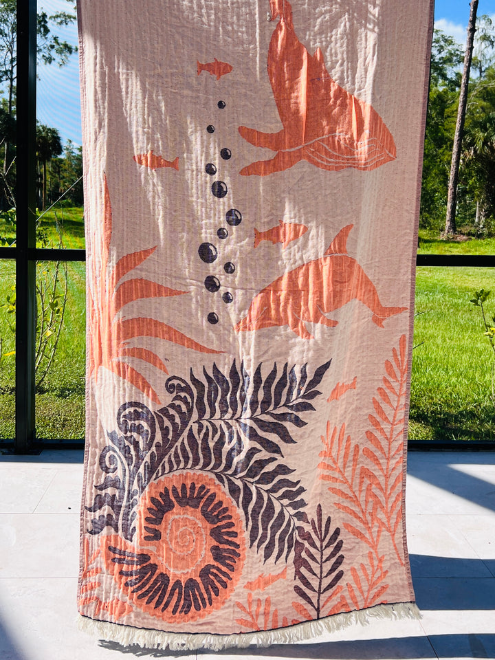 The Dolphin and Whale Turkish Towel by turkanhome.com, featuring beige jacquard weave with orange and dark blue sea life designs, hangs in sunlight with greenery visible through the window.