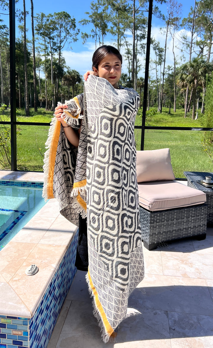 A woman stands by a poolside, wrapping herself in the Ikat Throw from turkanhome.com, featuring a reversible geometric pattern and yellow trim. Outdoor patio furniture and green trees are visible in the background.