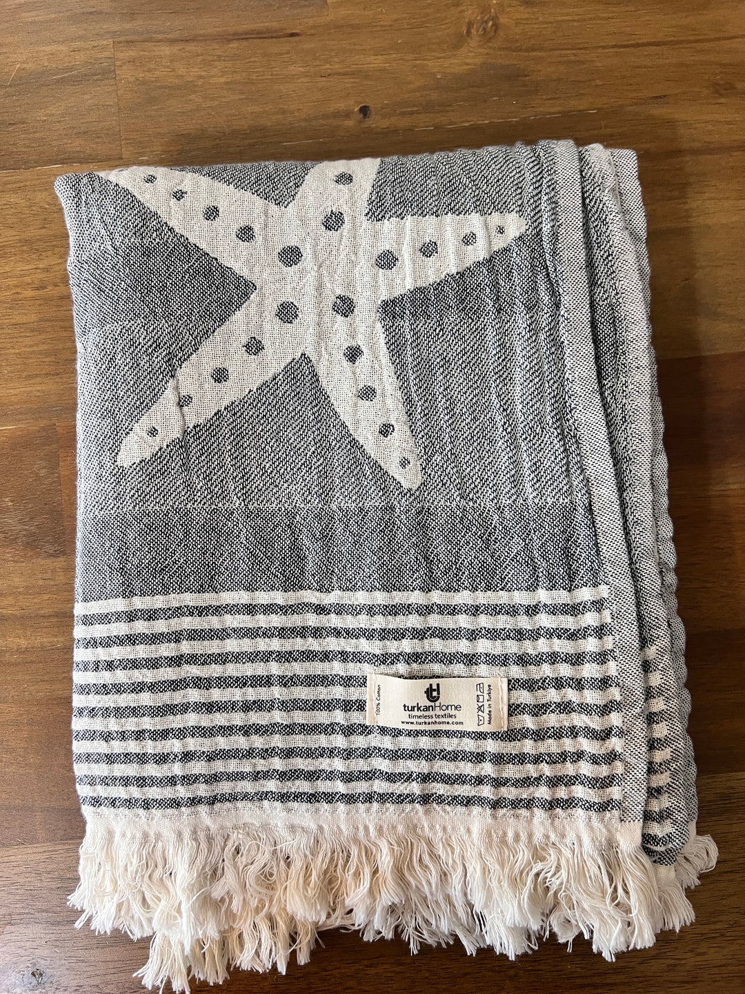 The Sea Star Turkish Towel from turkanhome.com, made of 100% cotton, features a gray and white Jacquard weave with a large starfish design, horizontal stripes, fringe on one end, and a small Turkish T label.