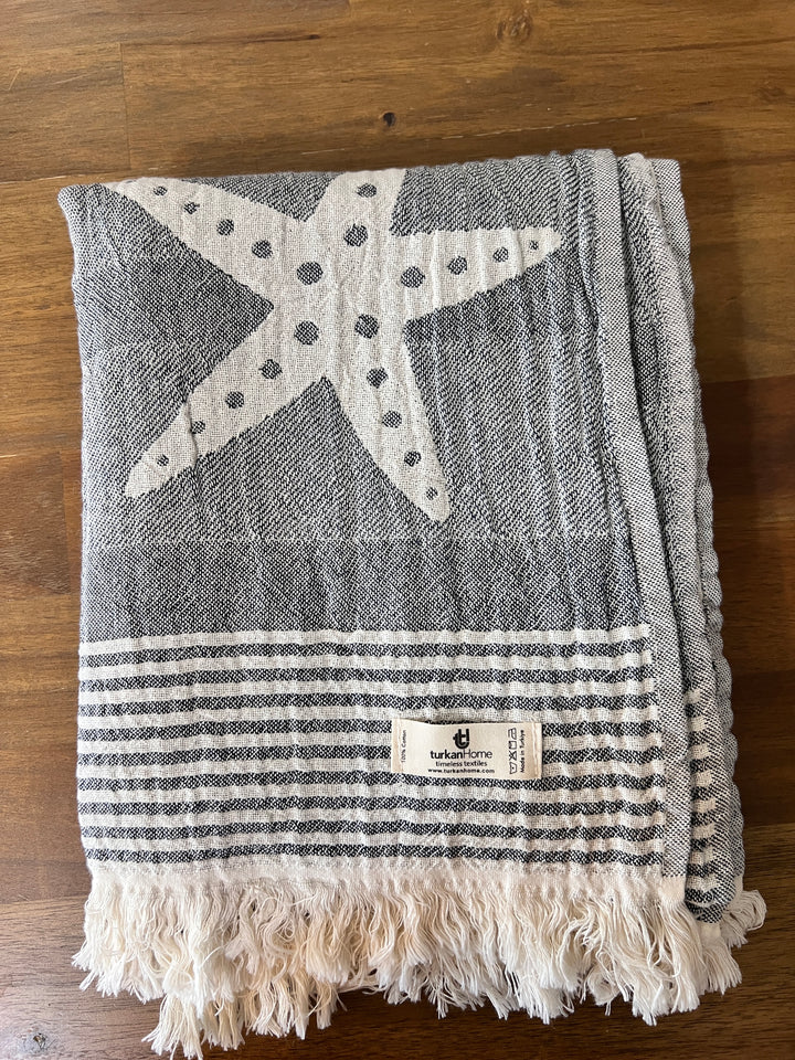 The Sea Star Turkish Towel from turkanhome.com, made of 100% cotton, features a gray and white Jacquard weave with a large starfish design, horizontal stripes, fringe on one end, and a small Turkish T label.