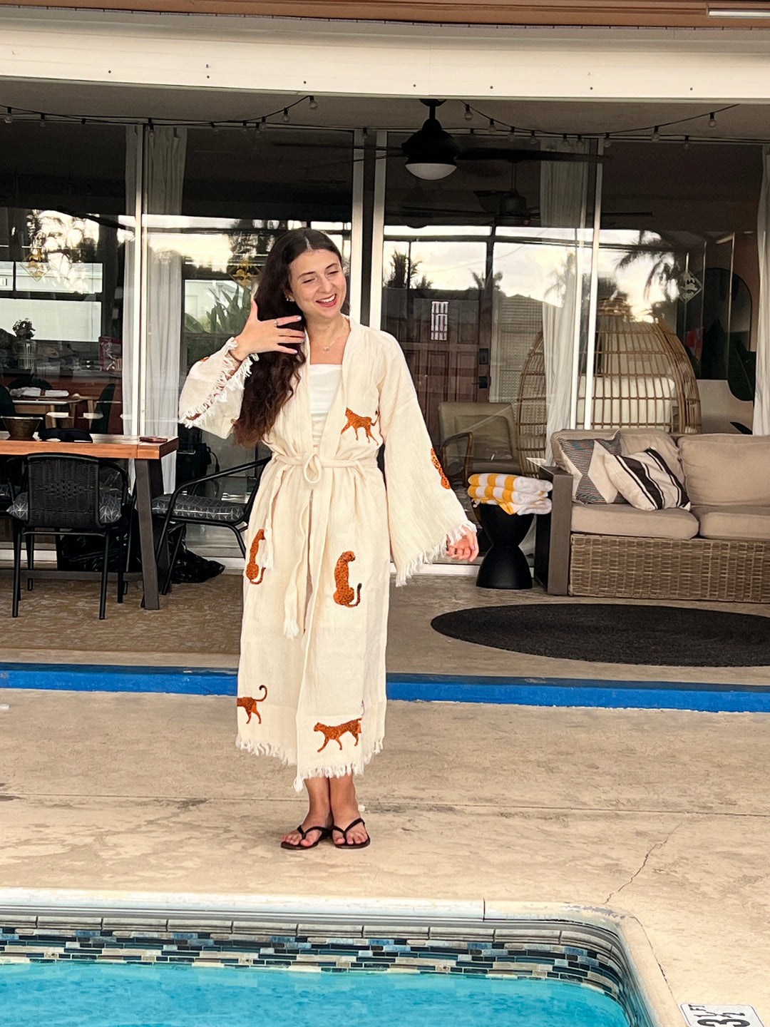 A woman stands poolside in a cream Orange Leopard Cotton Kimono Robe by turkanhome.com, holding her hair with one hand. Outdoor lounge furniture and a table are visible behind her.