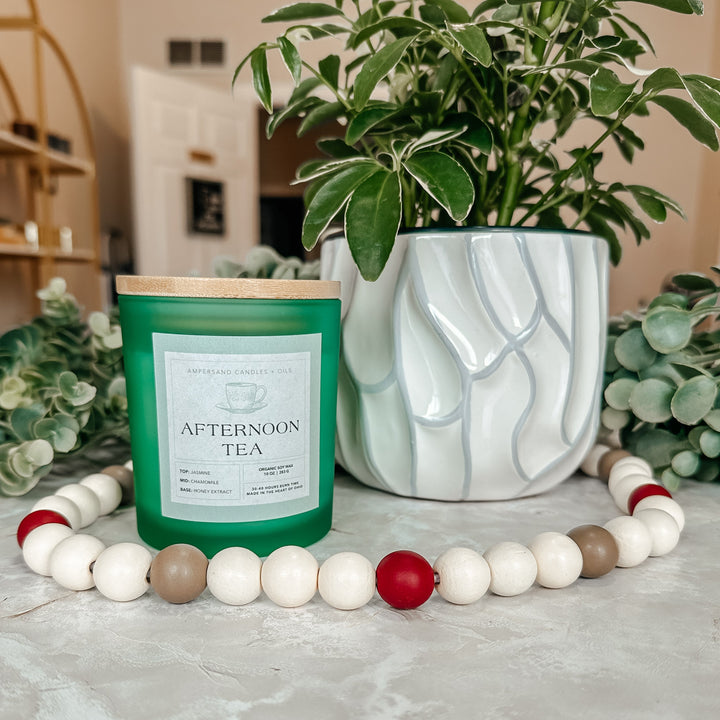 An Ampersand Candles + Oils Afternoon Tea Beeswax Candle (10oz) sits by a white plant pot with greenery, while a white, beige, and red bead garland in front adds charm to this natural candle display.