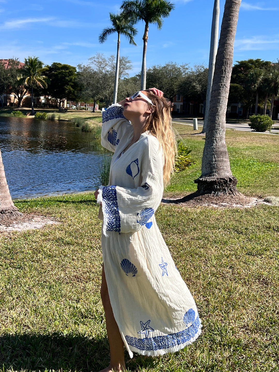 A woman wearing the turkanhome.com Sea Shell Kimono Robe-Blue with shell and starfish designs stands on grass near palm trees and a pond, looking up and holding sunglasses in bright sunlight.