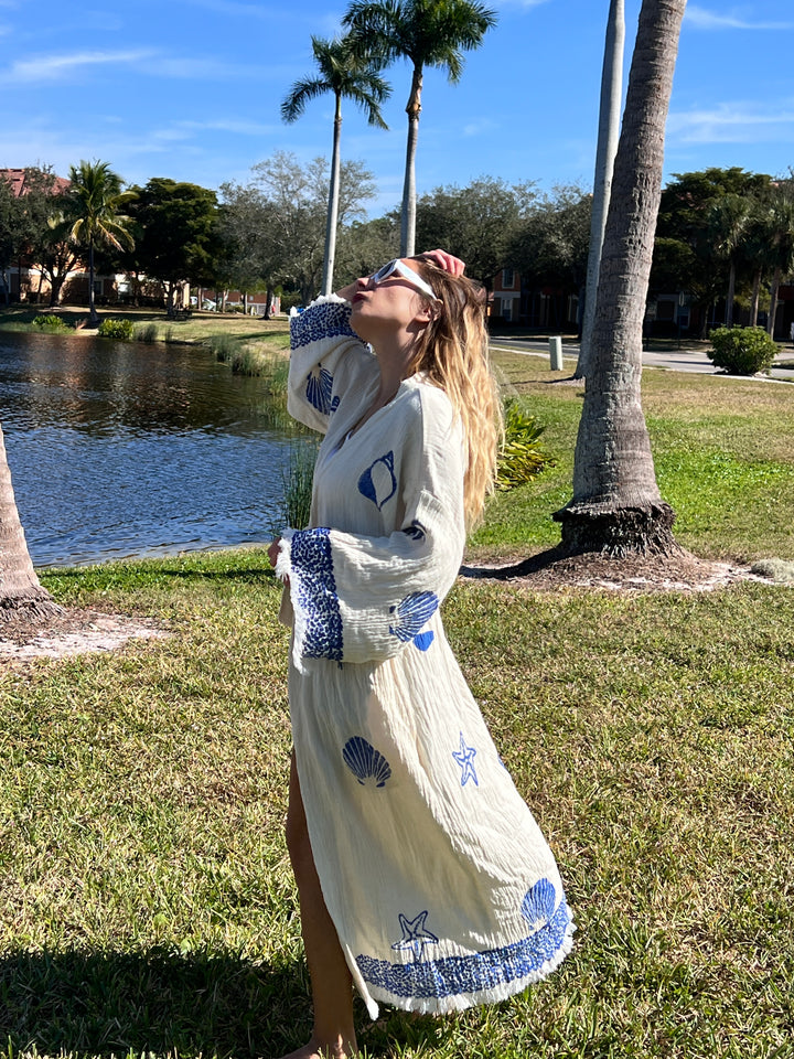 A woman wearing the turkanhome.com Sea Shell Kimono Robe-Blue with shell and starfish designs stands on grass near palm trees and a pond, looking up and holding sunglasses in bright sunlight.