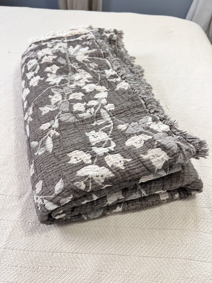 The Bougainvillea Muslin Bed Blanket – Brown | Queen by turkanhome.com, featuring a white floral pattern and fringed edges, is folded and resting on a light-textured surface.