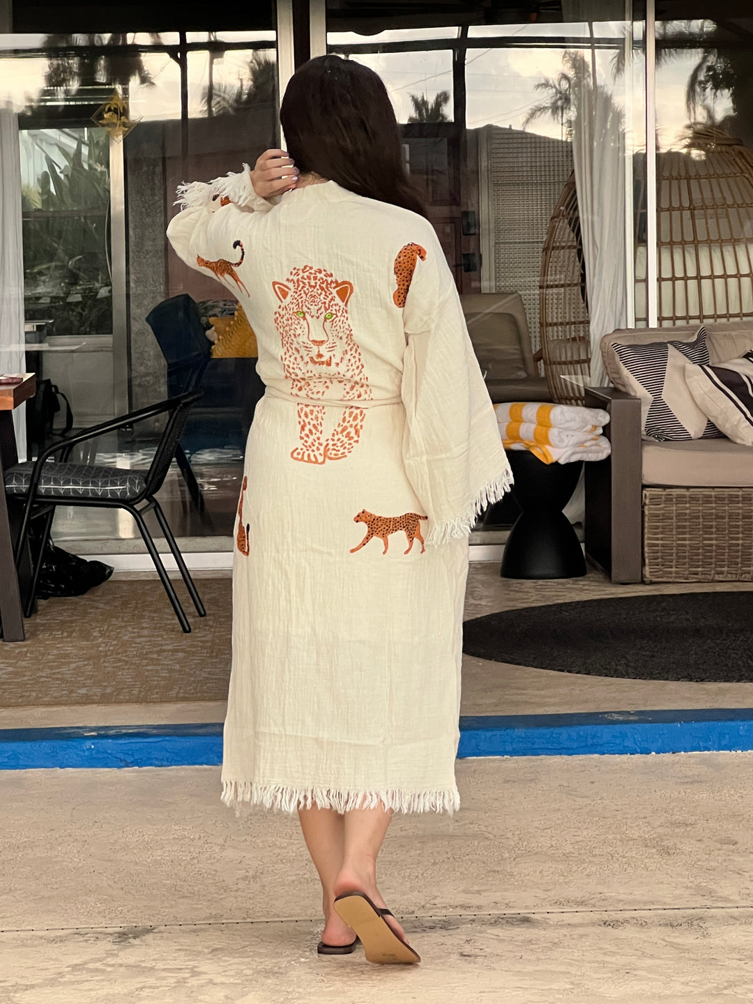 A woman stands poolside in the Orange Leopard Cotton Kimono Robe from turkanhome.com, a lightweight boho beach cover-up featuring orange and brown animal prints with a bold leopard on the back, surrounded by outdoor furniture.