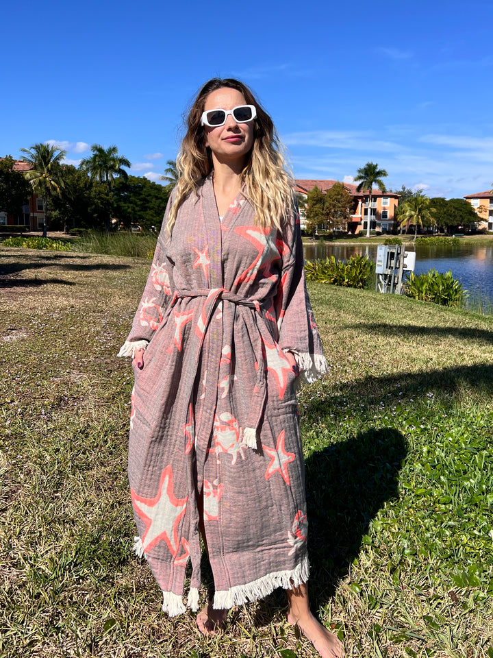 A woman in the Pink Sea Star Robe kimono by turkanhome.com and white sunglasses stands barefoot on grass by a lake, surrounded by palm trees and colorful buildings under a bright blue sky.