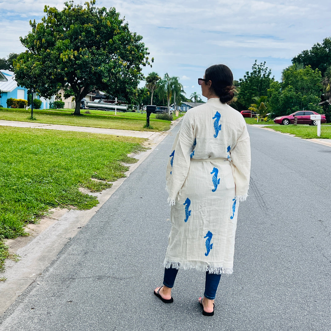 A woman faces away on a quiet suburban street, wearing the Seahorse Kimono Robe from turkanhome.com with blue seahorse prints, paired with jeans, sandals, and sunglasses. Green lawns and trees border the peaceful neighborhood.