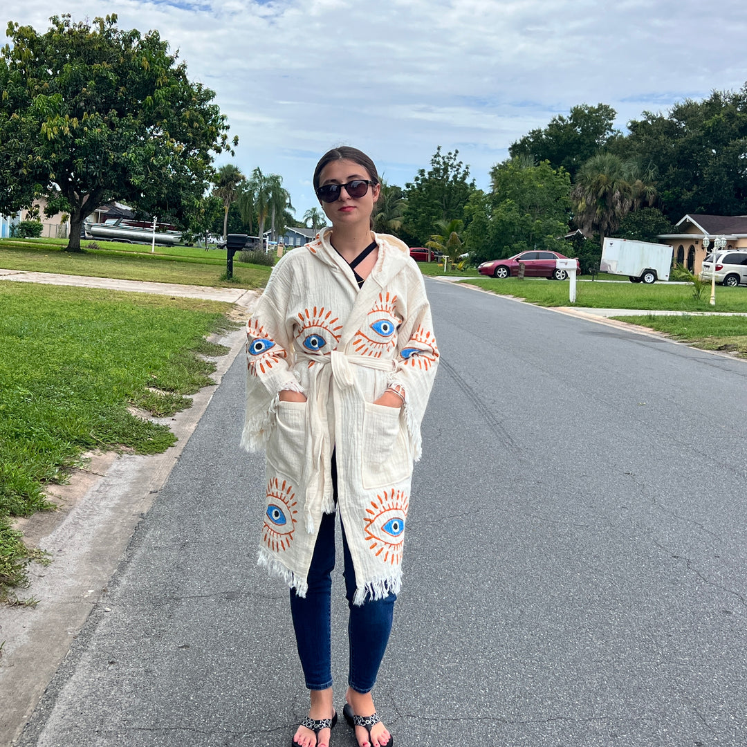 A woman stands on a suburban street wearing sunglasses, jeans, sandals, and the Mystic Eye Kimono Robe Short with Pockets and Hood from turkanhome.com. The cream-colored boho robe features blue and orange eye designs. Trees and houses are in the background.