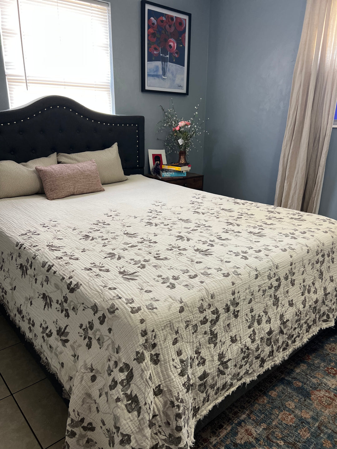 A neatly made bed features a white and gray floral quilt, beige and pink pillows, and the Bougainvillea Muslin Bed Blanket – Brown (Queen) from turkanhome.com, with a dark tufted headboard. A sunlit nightstand holds books, flowers, and a framed photo.