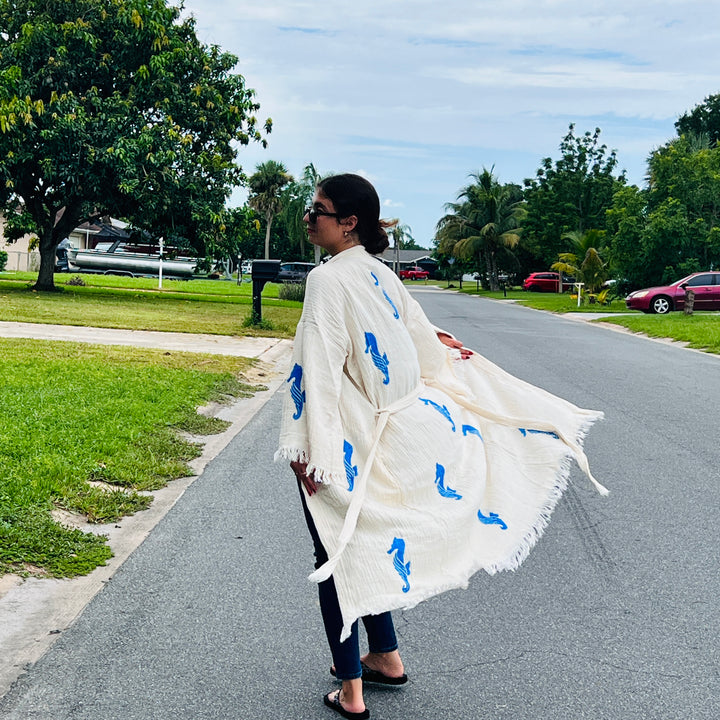 A woman in sunglasses and a long white Seahorse Kimono Robe with blue prints from turkanhome.com strolls along a tree-lined suburban street with lawns and parked cars under a partly cloudy sky.