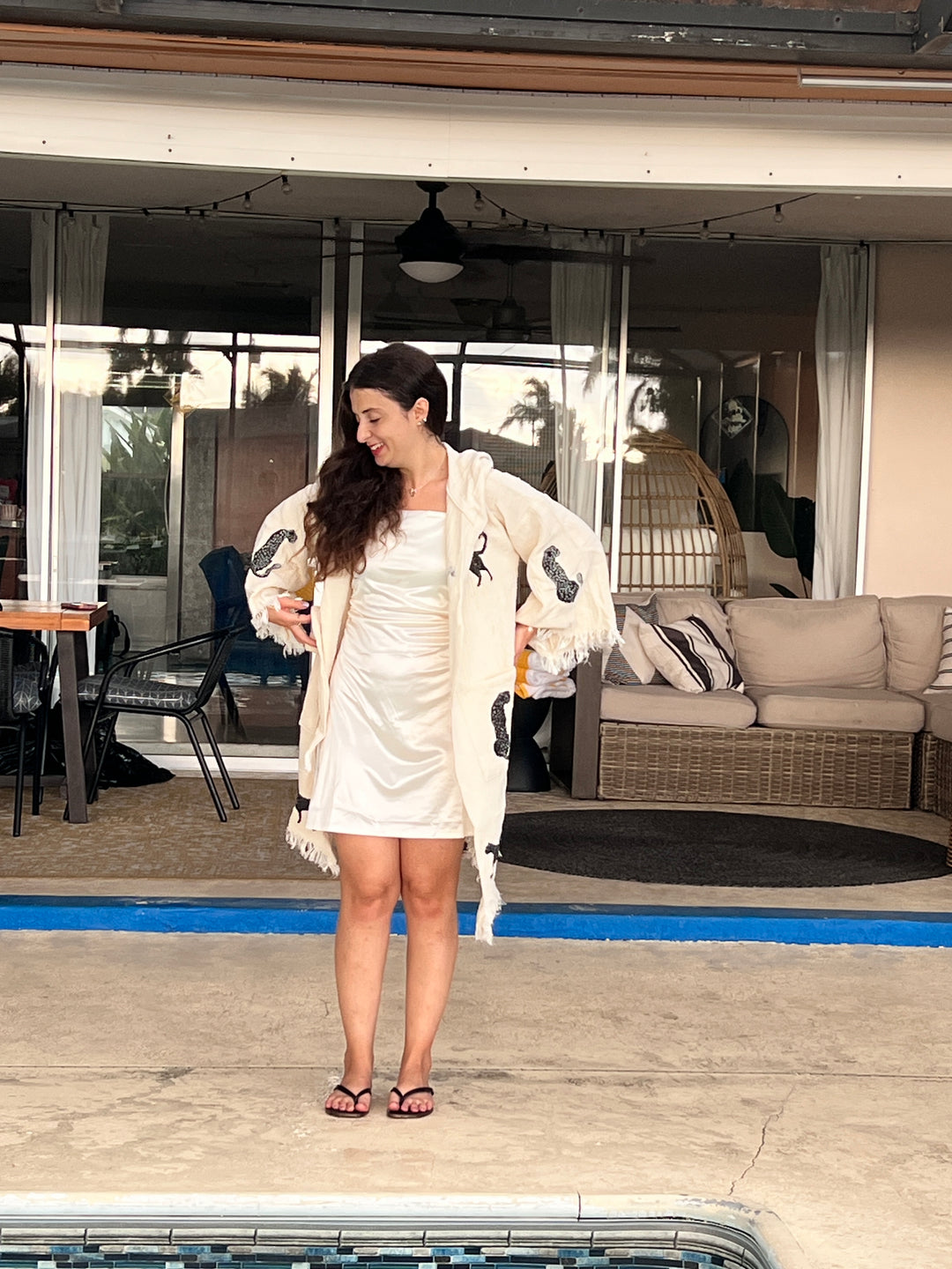 A woman in a white dress and Black Leopard Hooded Cotton Kimono Robe from turkanhome.com stands smiling near a pool, with outdoor furniture and large windows in the background.